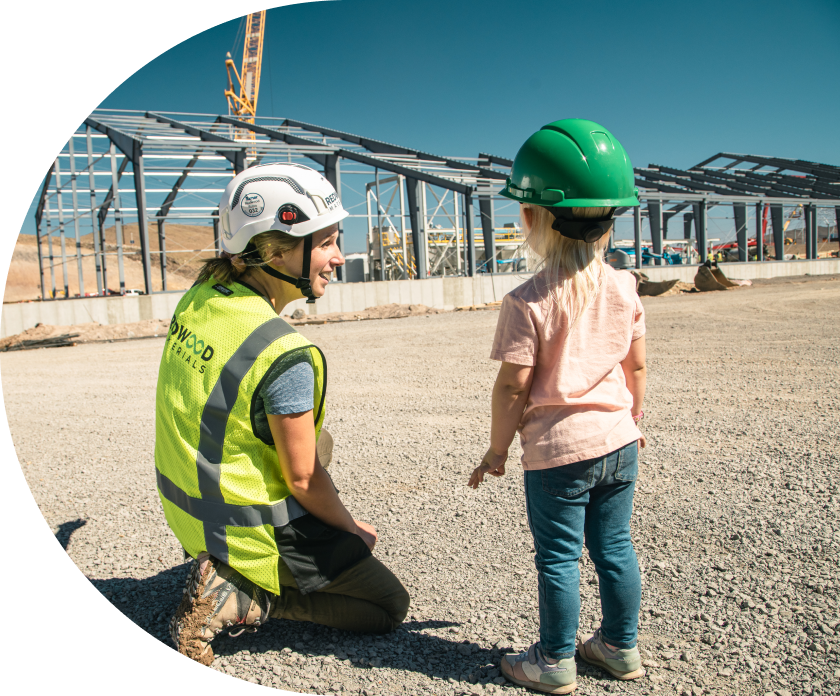 Worker in hard hat and vest kneeling and showing construction site to a small child