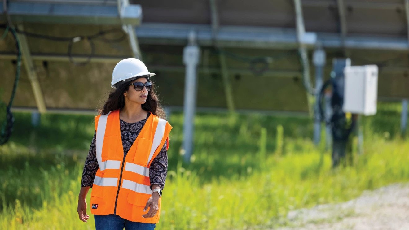 A woman wears an orange safey vest, a hard hat, and sungalsses as she walks outside by a solar farm